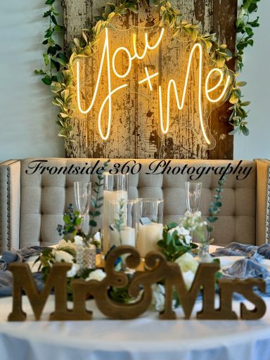"Wedding decor featuring 'You & Me' neon sign and 'Mr & Mrs' wooden sign on a table."