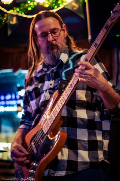 Man playing an electric bass guitar wearing a plaid shirt in a dimly lit setting.
