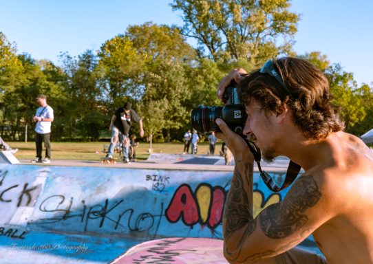 A shirtless photographer captures a skateboarding scene at a park.