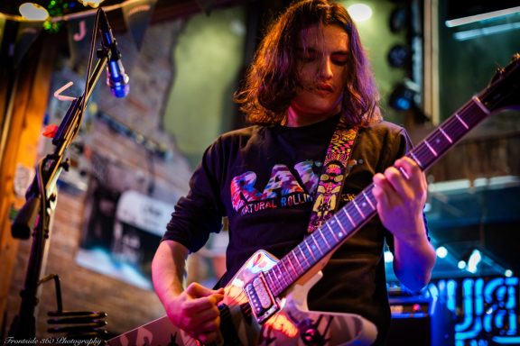 Musician playing guitar on stage with colorful lights and a lively atmosphere.