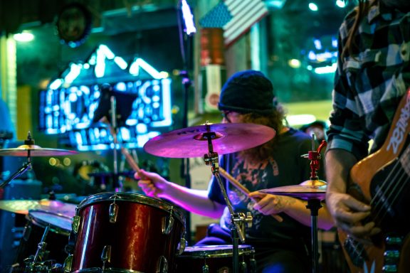 Drummer performing on stage with colorful lights and a band member in the foreground.