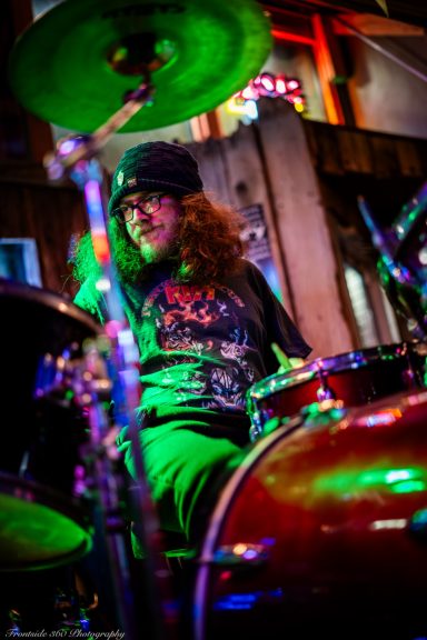 Drummer with curly hair wearing a beanie, playing in a vibrant, colorful setting.