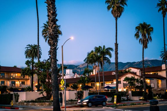 Street scene at dusk featuring palm trees, buildings, and distant mountains.
